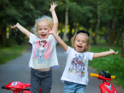 Two Little Girls Standing Behind Toy Scooters and Raising Hands