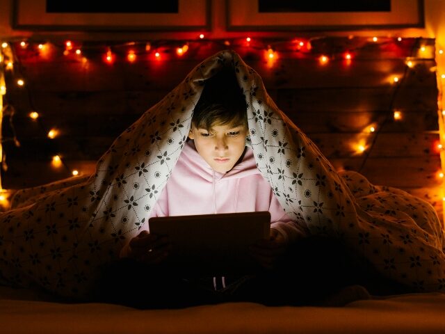 Boy lying on bed under blanket and using a tablet