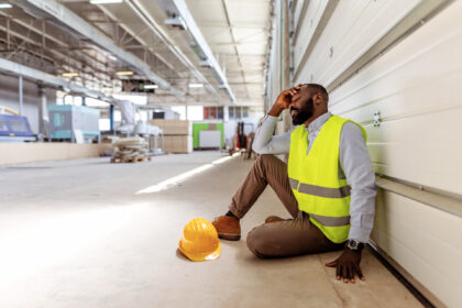 Worker in a high-visibility vest seated in a large industrial warehouse, deep in thought,