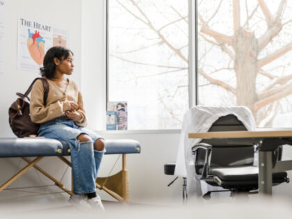 The worried and anxious young adult female student looks out window of the doctor's examin