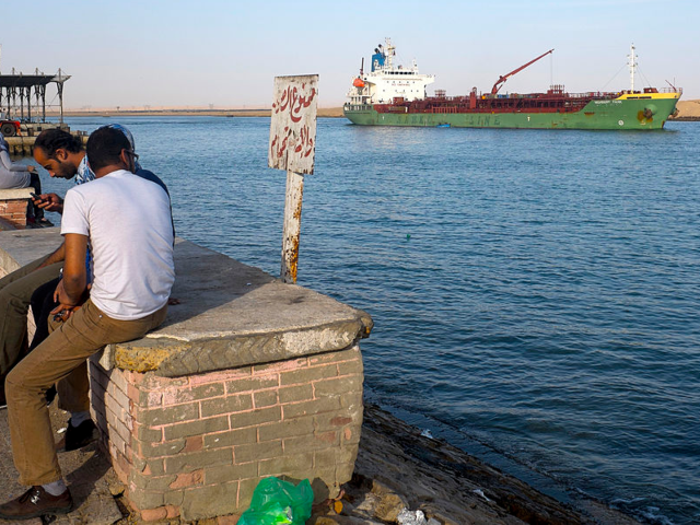 GettyImages-166406220 People sit and watch commercial ships as they pass through the entrance to the Suez Canal