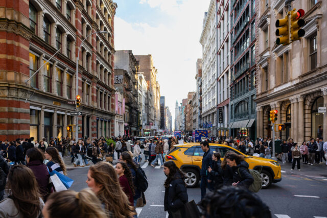 Many people are walking across the street in Manhattan New York City with a yellow taxi ca