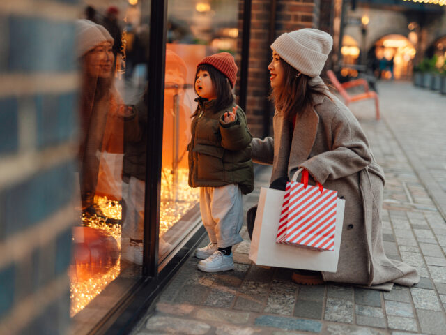 Curious Asian toddler girl looking at window display while doing Christmas shopping with h
