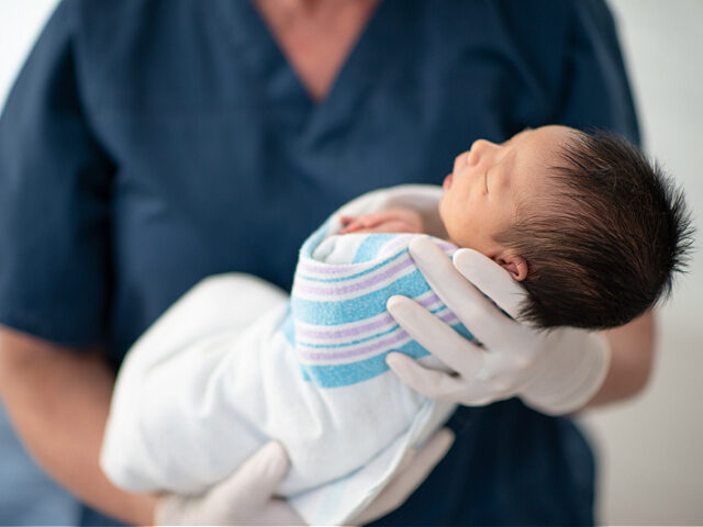 A female nurse of is holding a newborn baby at the hospital. They baby is sleeping.