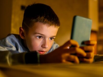 GettyImages-2214023900 Young boy attentively interacts with a smartphone while lying indoors, illuminated by soft