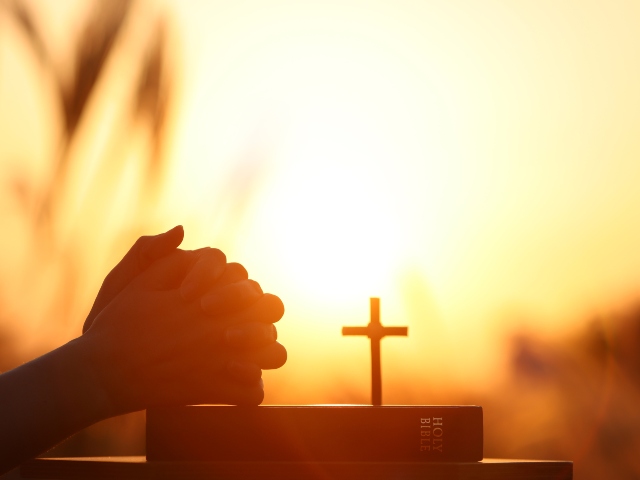Christian holding a Bible and praying with hands clasped together over an open cross