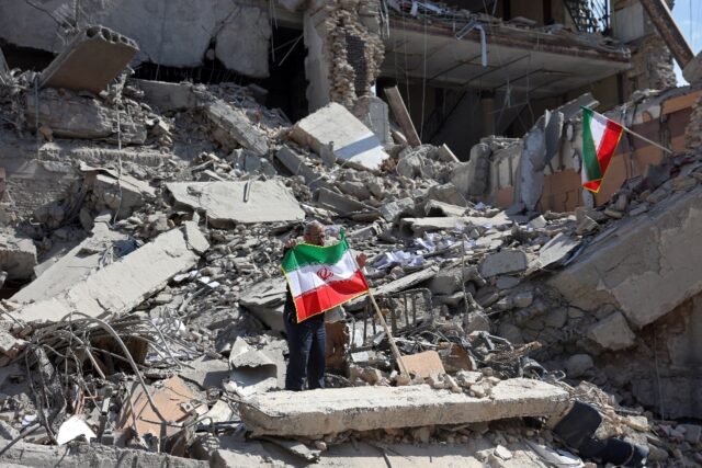 A man holds an Iranian flag amid the debris of a destroyed building following airstrikes i