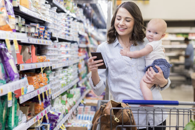 Working mom holds her adorable baby girl while using a smart phone in a grocery store. She