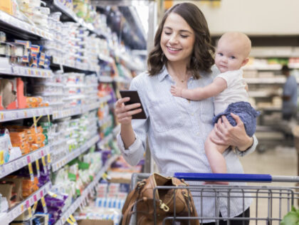 Working mom holds her adorable baby girl while using a smart phone in a grocery store. She