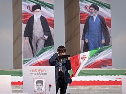 An Iranian schoolboy salutes while standing beside a portrait of a child killed in an airs