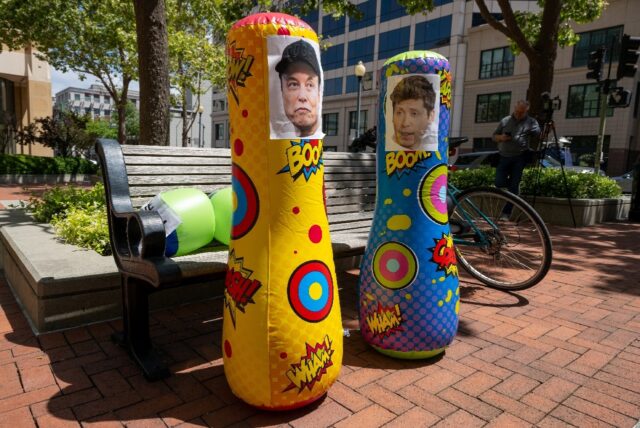 Punching bags with the faces of AI founders Elon Musk and Sam Altman are placed in front o