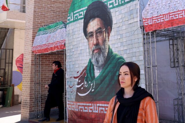 A woman walks past a large tiled image of Iran's current leader Ayatollah Mojtaba Kha