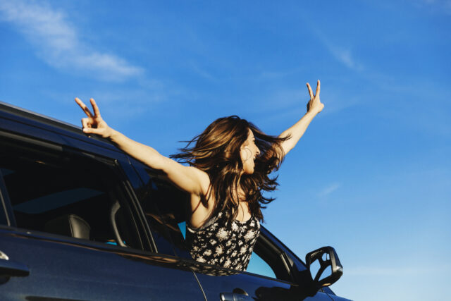 Carefree woman leaning out from car window with arms outstretched