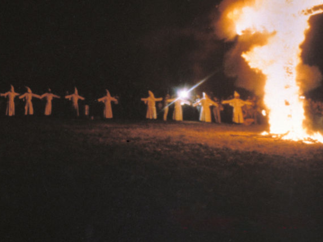 Ku Klux Klan members, in white robes and hoods, gather around a burning cross during a nig