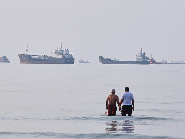 Two men wade in the waters of the Strait of Hormuz with vessels anchored in the background, off Bandar Abbas, Iran, Sunday, July 12, 2026. (Razieh Poudat/ISNA via AP)