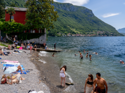 Sunbathers and swimmers are seen at a lakeside beach with red and yellow buildings on the