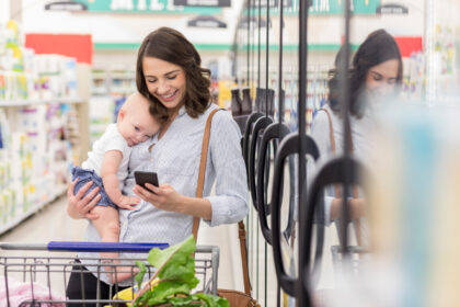 Happy young Hispanic mother texts while holding baby girl in a grocery store. She is shopp