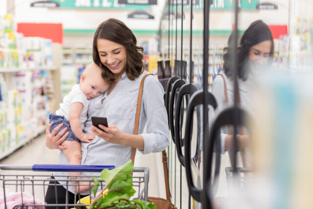 Happy young Hispanic mother texts while holding baby girl in a grocery store. She is shopp