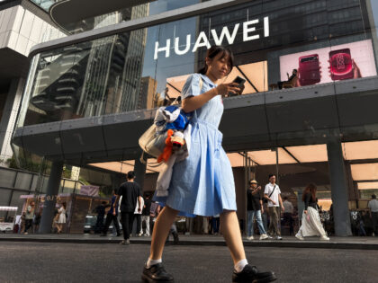 A pedestrian looks at a smartphone while walking past a Huawei retail store displaying an