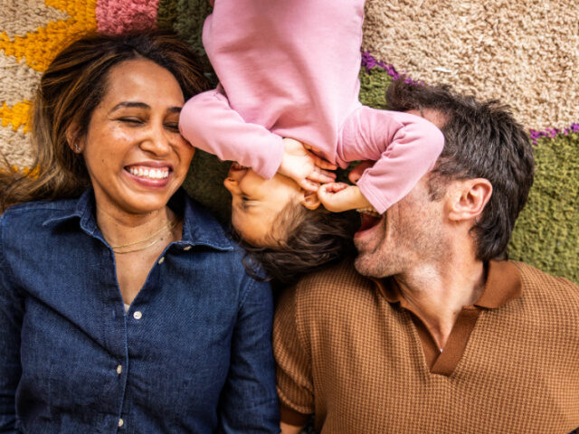 Mother and father laughing with toddler daughter while laying on rug