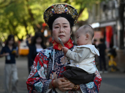 A woman dressed in imperial costume holds a baby as it plays with a mobile phone in a stre